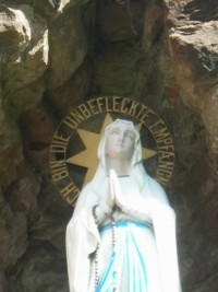 La statue de Notre-Dame de Lourdes est surmontée d'une auréole dorée, portant les paroles par lesquelles Elle s'est présentée à sainte Bernadette à Lourdes.
